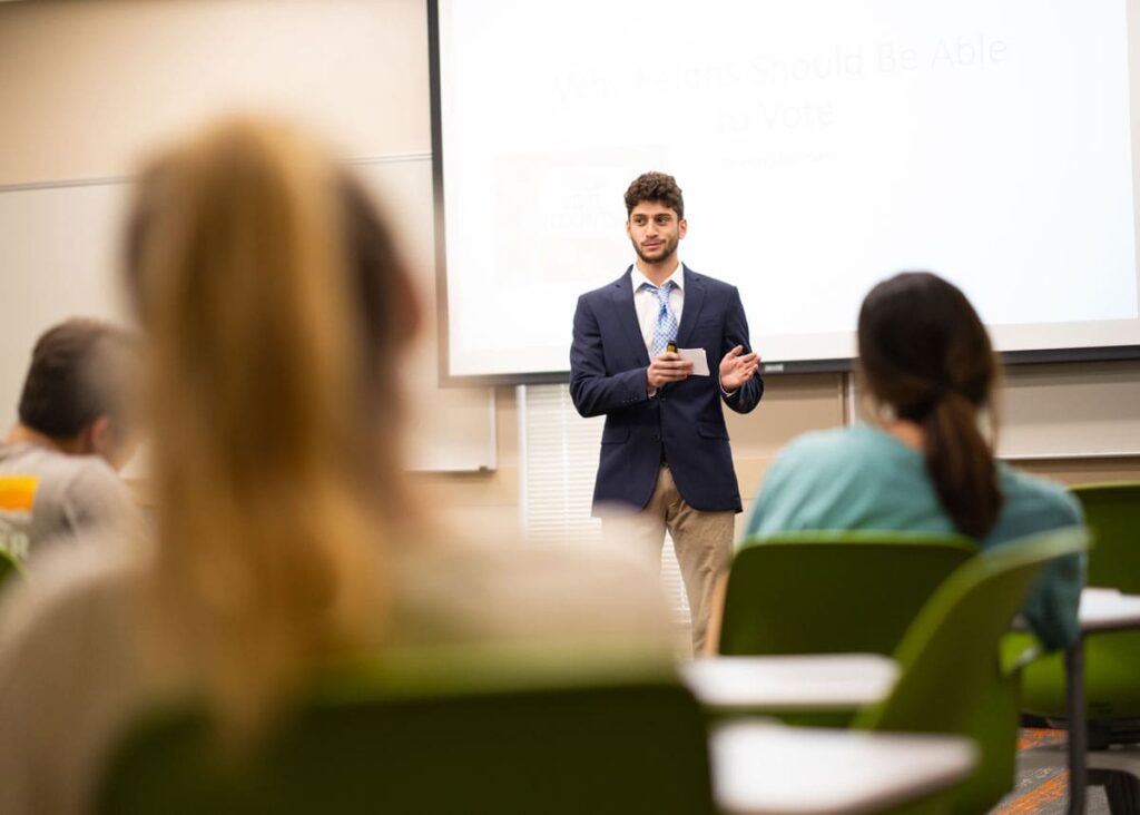 A student wearing a business casual outfit gives a speech at the front of a room in front of other students.
