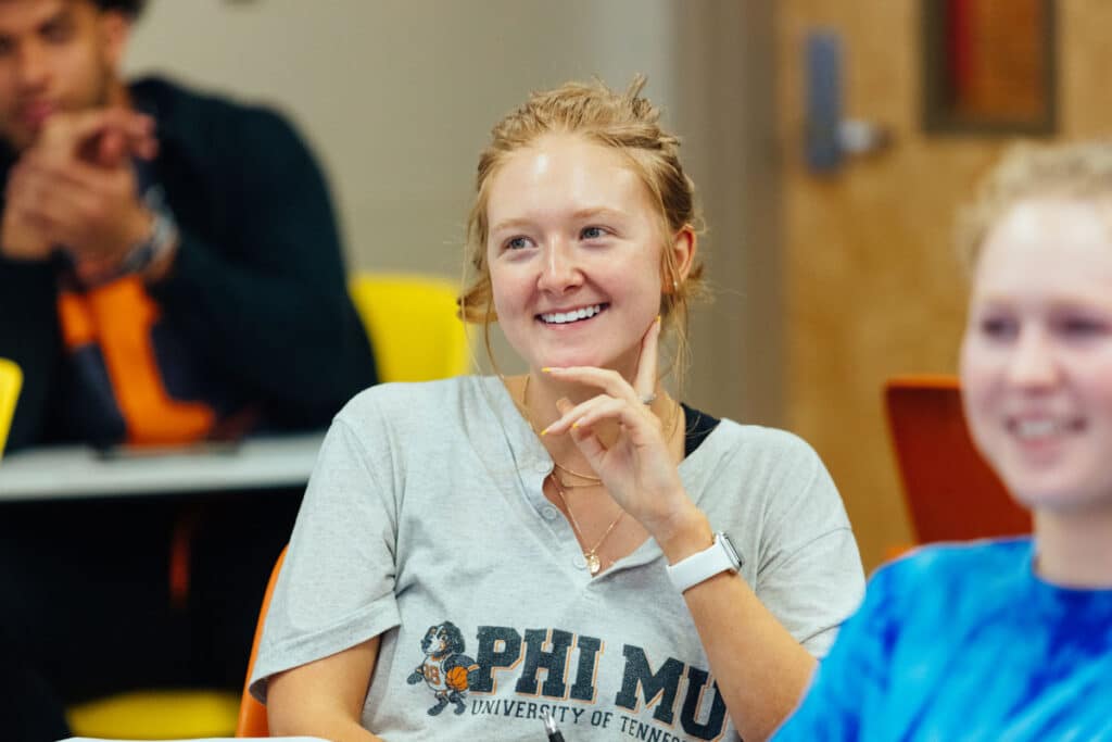 A student holds her left hand against her face and laughs while seated in a class with other students in the foreground and background.