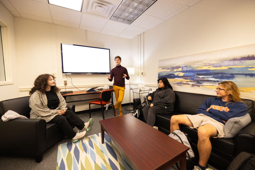 A student stands in front of a TV screen and talks to three other students seated on couches in the Message Effects Lab.