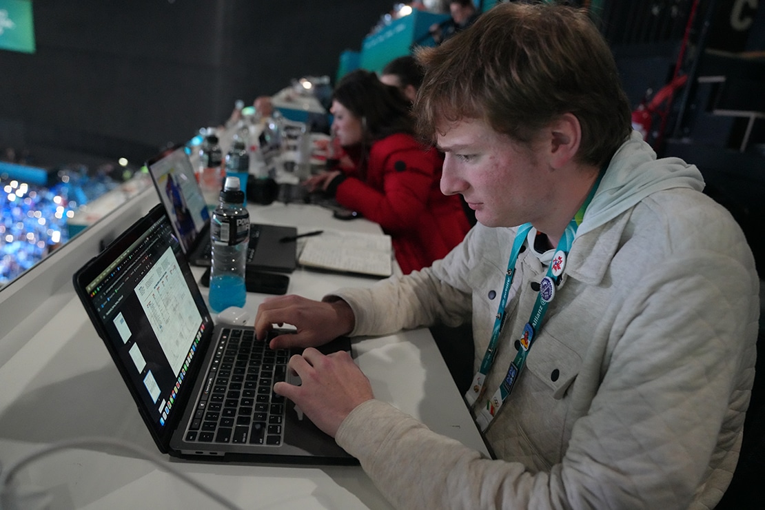 A person in a white jacket focuses intently while typing on a laptop in a bustling sport arena setting. He's wearing a lanyard with several pins on it and has blonde hair. There are people blurred in the background.