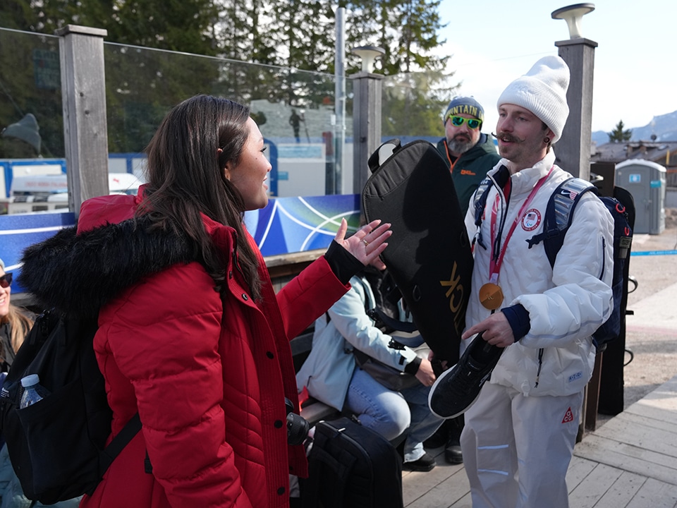 A smiling man in a white winter outfit, holding a snowboard and wearing a medal, talks to a woman in a red coat outside, with mountains in the background.