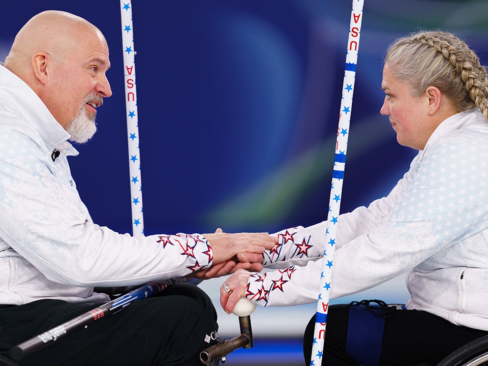 Two wheelchair curlers in white jackets with star patterns face each other, holding hands. The scene conveys teamwork and support. A calm and focused atmosphere.