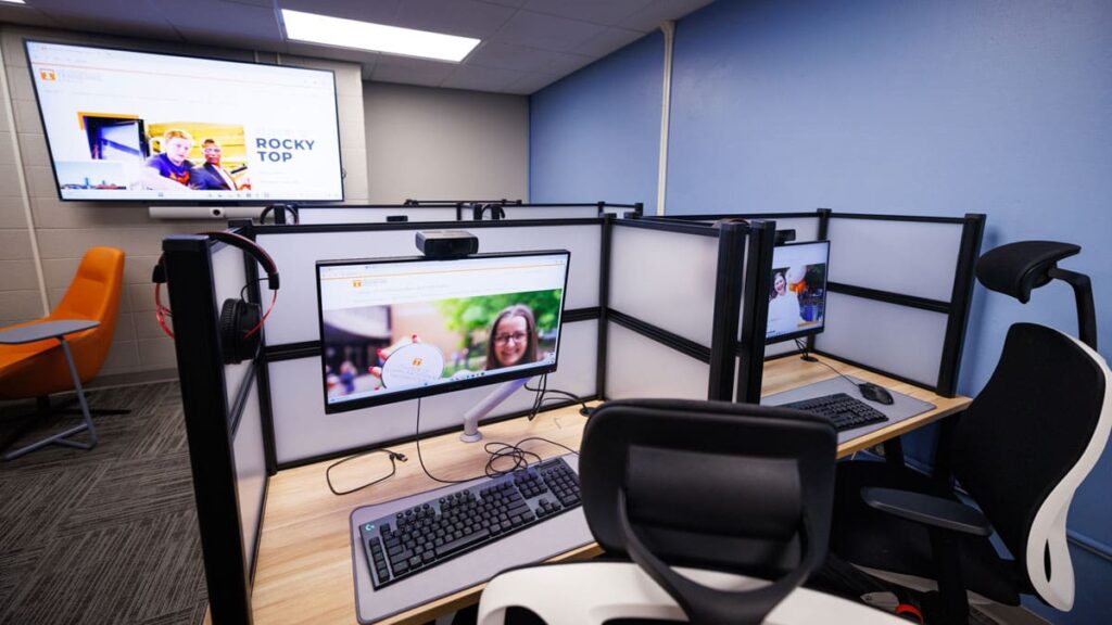 The iLab is show with cubicles featuring two rows of cubicles, two to a row, each with computers, keyboards, and a nice desk chair. In front of the cubicles on the far wall is a large screen.