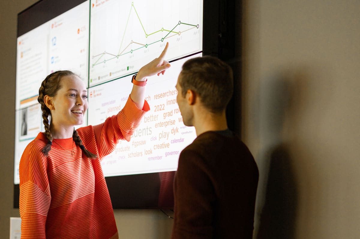 A student in an orange sweater points toward the screen in the Adam Brown Social Media Command Center as she talks to another student standing in front of her.
