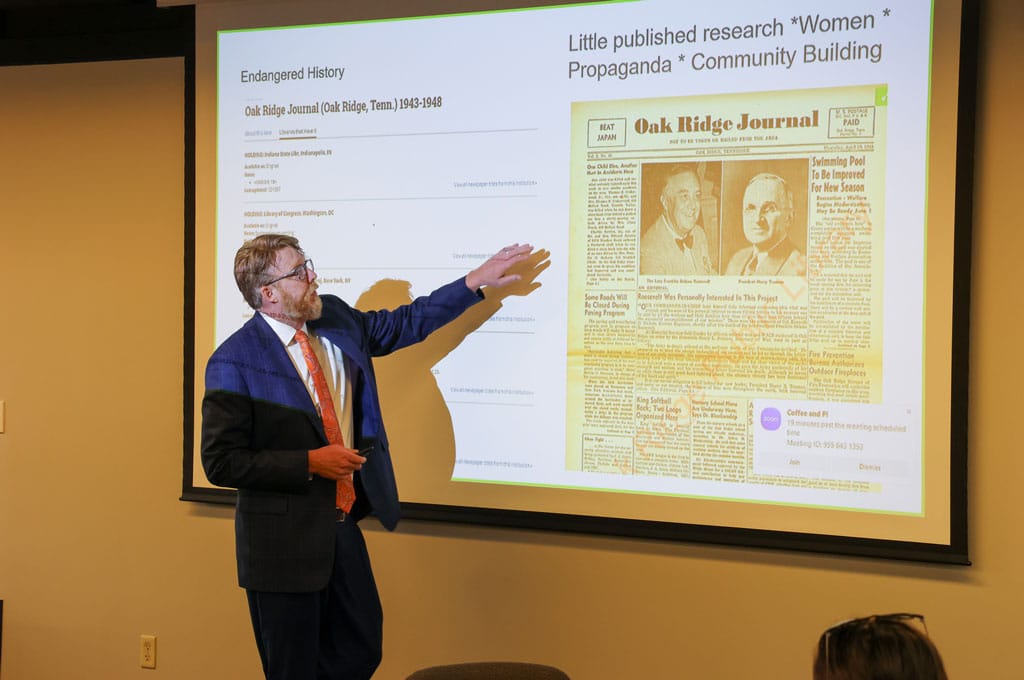 A man wearing a blue suit and orange tie gestures towards a presentation on a large screen behind him