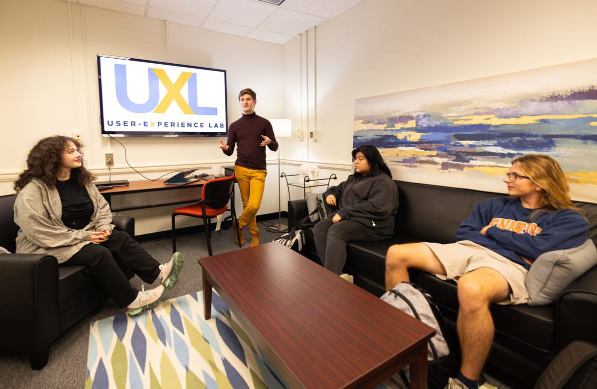 A student gestures with both arms as he stands by a table in front of a large TV screen on the wall and three students on couches around him listen. They are in the Message Effects Lab, the tv screen says U-X-L.