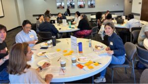 Students and faculty sit at several tables in a room while eating breakfast.