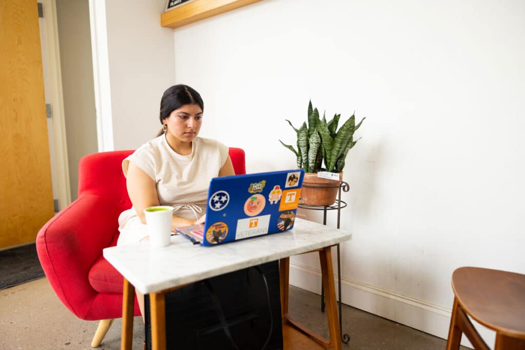 A nontraditional student sits at home at a white desk, in a red chair, working on her laptop.