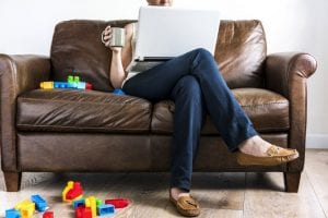 A parent sitting on a couch with a laptop and coffee with legos on the floor and couch