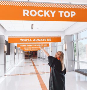 CCI alumna Kylie Julius in her master's gown makes a peace sign as she walks the hallway in the Student Union with an orange and white banner that reads "Rocky Top" above her.