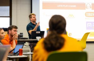 Tombras ADPR Assistant Professor Matthew Pittman gestures as he teaches students. A slide behind him says "Let's play a game."