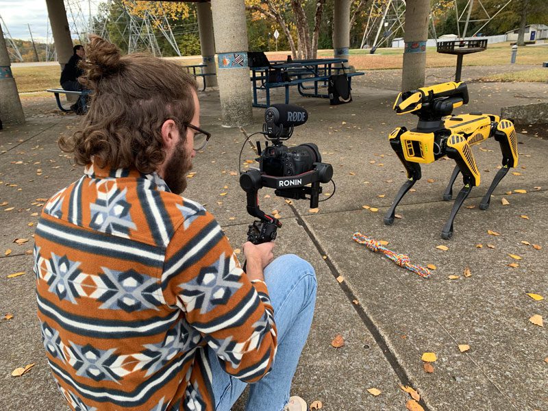 A student in a colorful sweater uses a video camera to record a black and yellow dog robot for a JobPop piece for PBS.