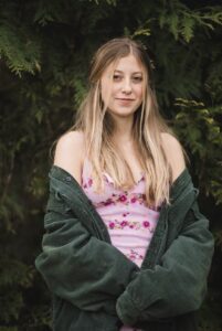 Ava Franzoy wears a pink floral spaghetti strap dress with a large dark green corduroy jacket hanging off her shoulders, her hands held in front of her, while standing in front of dark green foliage.
