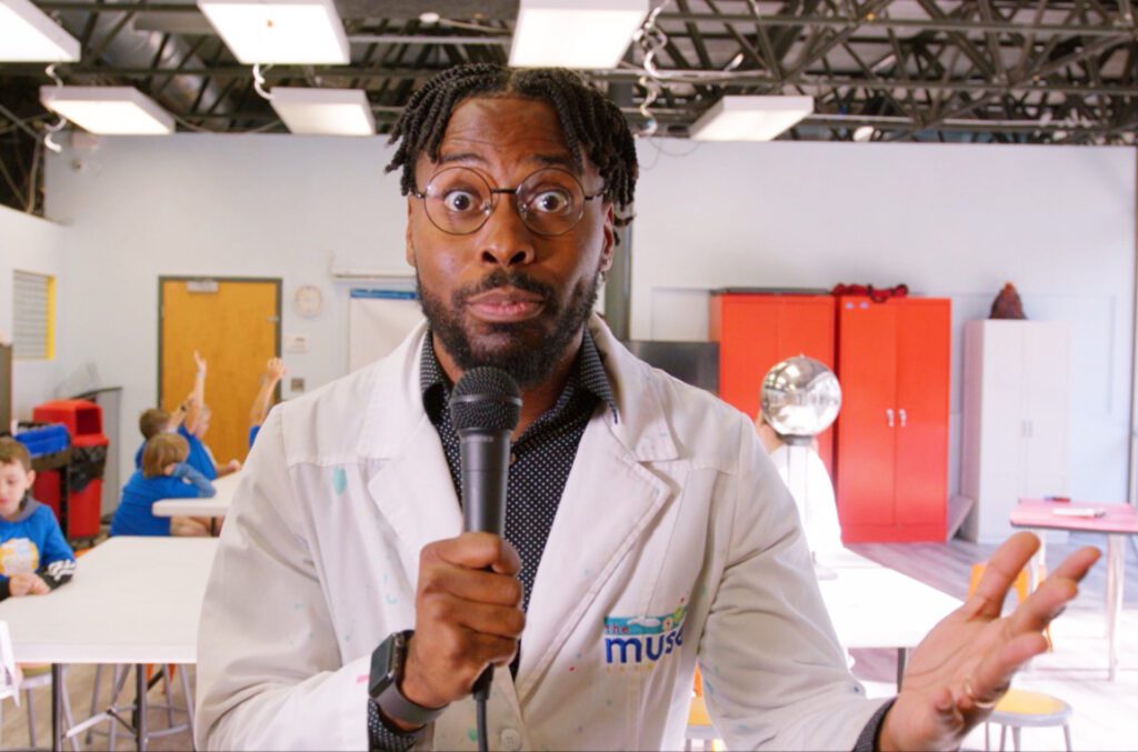 Alumnus Tyrone Beach dressed in a scientist coat and holding a microphone at his job at Muse Knoxville.