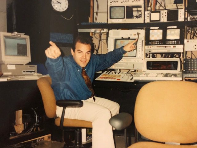 A young Barry Rice holds his arms out in celebration while sitting in a video production office.