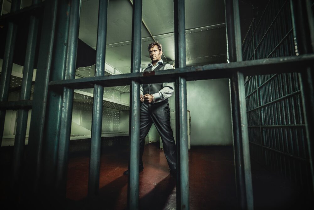 A stylized picture of Curt Anderson with a suit and handcuffs on, viewed through jail cell bars, as he prepares to do a magic trick.