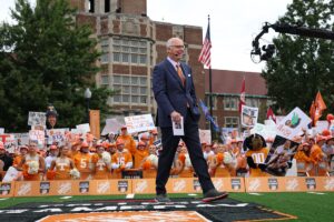 Gene Wojciechowski wears a suit and orange and white tie as he walks onto the Neyland Stadium football field by a corridor of football fans decked out in Tennessee organe and holding up signs.