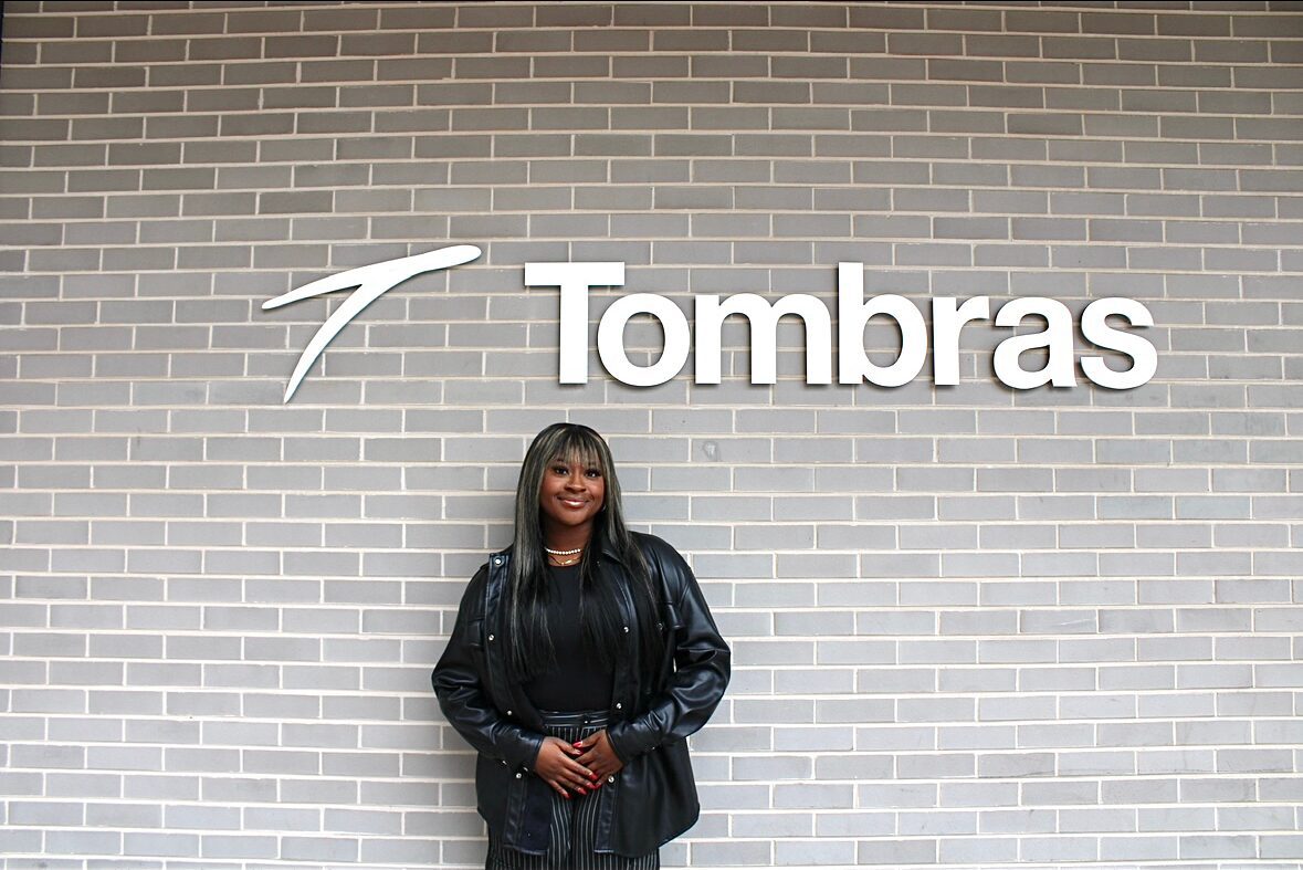 Senior Faith Mtabo stands in front of a grey wall with the Tombras logo above her.