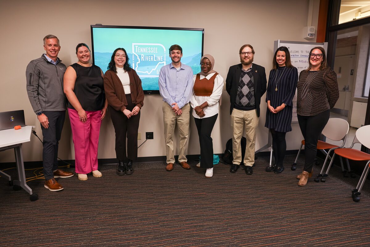 A group of eight graduate students and professors stand in the Adam Brown Social Media Command Center in a row in front of a large presentation screen.