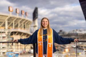 Mackenzie Ostrom is dressed in University of Tennessee, Knoxville, graduation garb and holding her arms out wide to her sides with Neyland Stadium in the bakground.