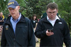 John Tupps stands next to then-Florida Governor Rick Scott, talking and looking at a phone.