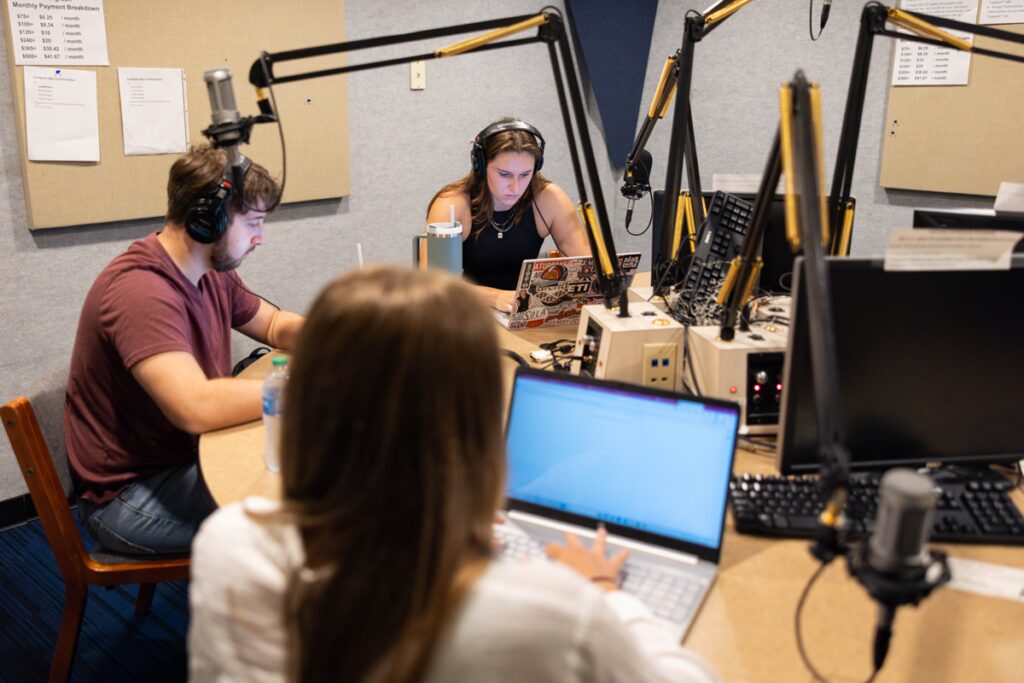 Three graduate students, Katie Mack, Nick McNeese, and MaryBeth Mahne, sit around a circular desk at WUOT that has microphones positioned above them. They are all deeply engrossed in their computers and the conversation.