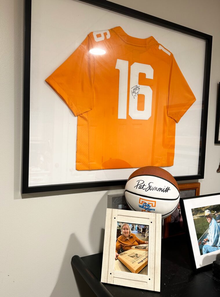 An orange and white Tennessee Jersey with the number 16 is displayed in a frame on the wall, and below it sits a basketball signed by Pat Summitt, next to a picture of Linda Higgins looking at archival copies of the Orange and White newspaper.