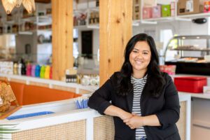 Anna Myint, in a black suit jacket and white and black striped shirt, poses at the countertop of her family's restaurant International Market.