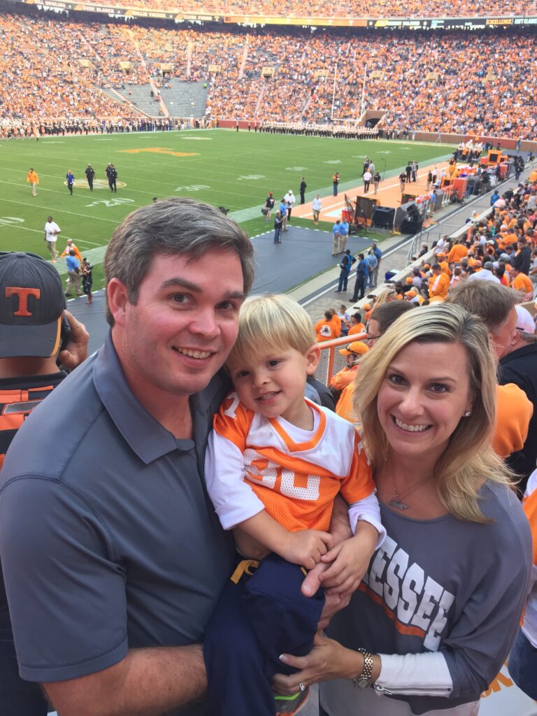 Lee and Erin Freeman pose in Neyland Stadium with the football field in the background and their young son, John Clark, held between them.