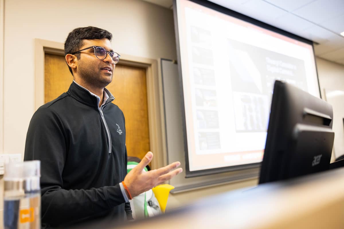 PhD student Aman Misra stands behind a podium with a computer monitor on it and a projector screen to his left while teaching a class.