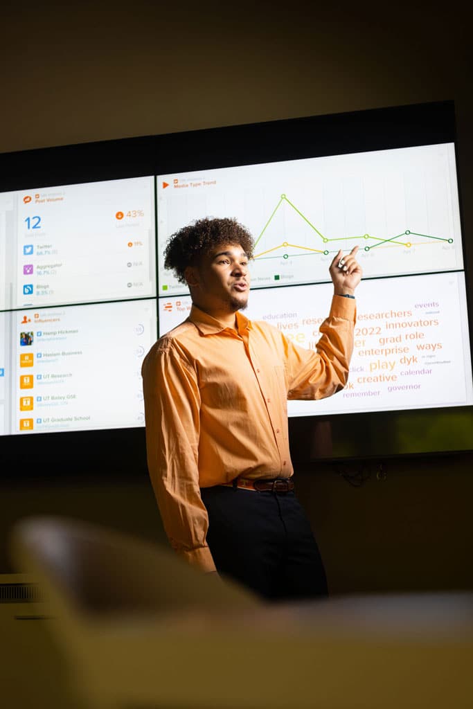 A student wearing a UT orange long-sleeved shirt is giving a presentation in front of a large screen displaying social media analytics.