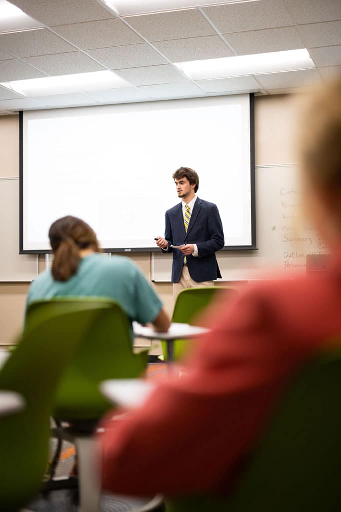 A student in business professional clothing stands in front of a white projector screen and gives a class presentation.