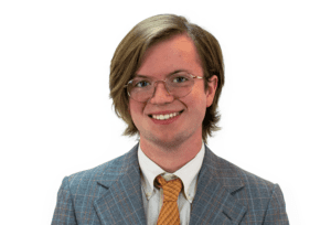 A headshot of Pierce Gentry wearing a grey, orange, and baby blue plaid suit jacket, white shirt, and orange tie.