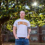 Jonathan Gilman stands with hands in the pockets of his medium wash jeans, wearing a white T-shirt with Tennessee written across the chest in UT orange, trees and a brick campus building behind him.