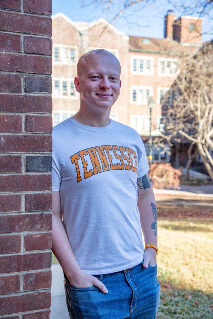 Jonathan Gilman leans against a brick wall on the UT campus while wearing a white shirt with the word Tennessee written across it in UT orange.