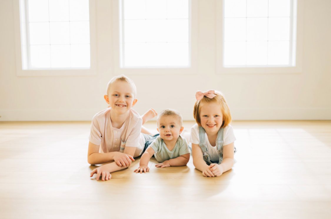 Three smiling young Rowbury children lay on their bellies on the floor facing the camera for a professional portrait.