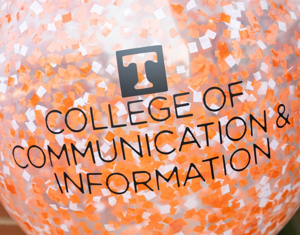 A close-up of a balloon with orange and white confetti in it and the College of Communication and Information logo on it in black.