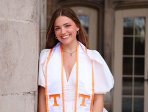 Skylar Brown leans against a column on the UT campus and wears a crisp white dress with a v neckline and poofy sleeves with her UT stole around her neck.