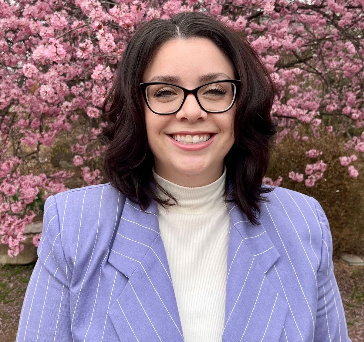 Tori Bertram headshot of her wearing a white turtleneck with a lavender suit coat with wide-set white pin stripes and in the background behind her is a blooming cherry tree.