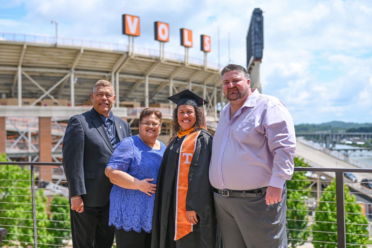 Melissa Espinales wears her graduation regalia and stands with, from left, her father, mother, and husband, on the College of Communication patio with Neyland Stadium and the Vols sign in the background behind them.