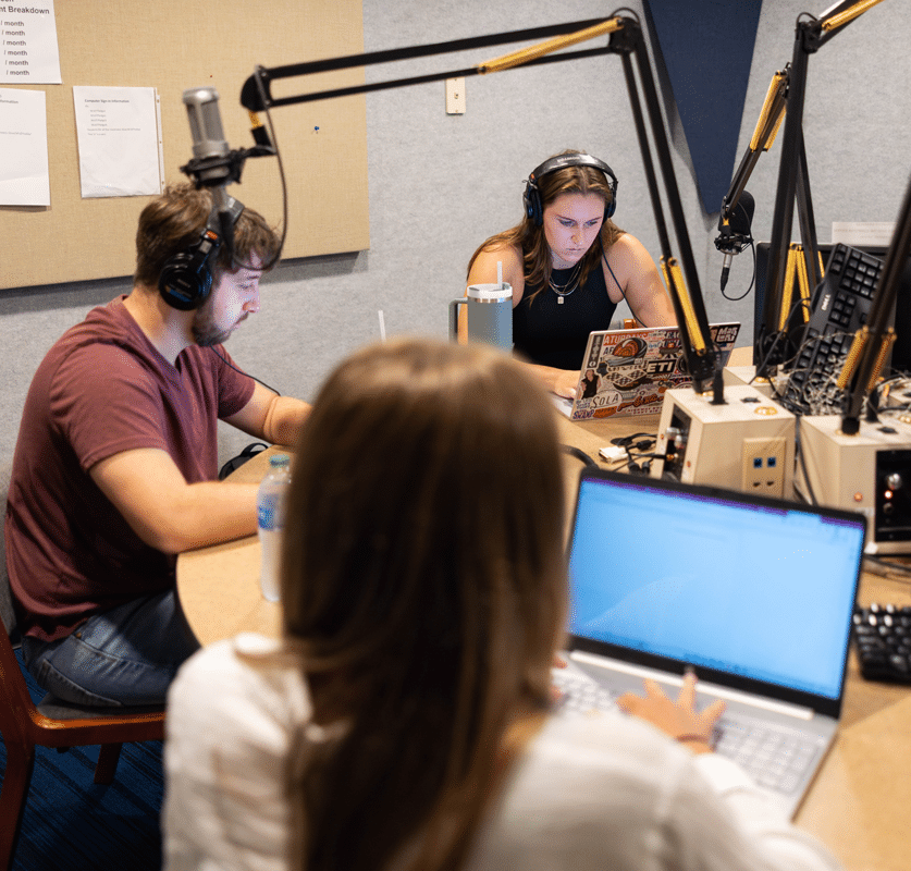 Three students sit in a WUOT studio, all looking at their laptops and two wearing headphones.