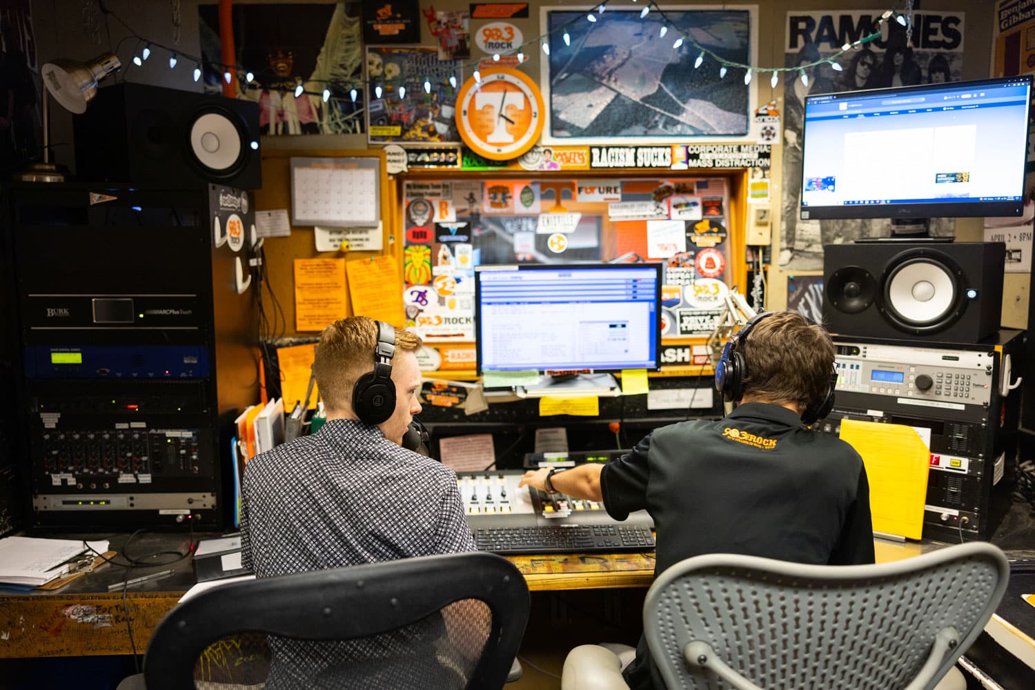 two students sit in front of a computer screen and a wall full of WUTK and music posters and memorabilia inside of a WUTK station studio.