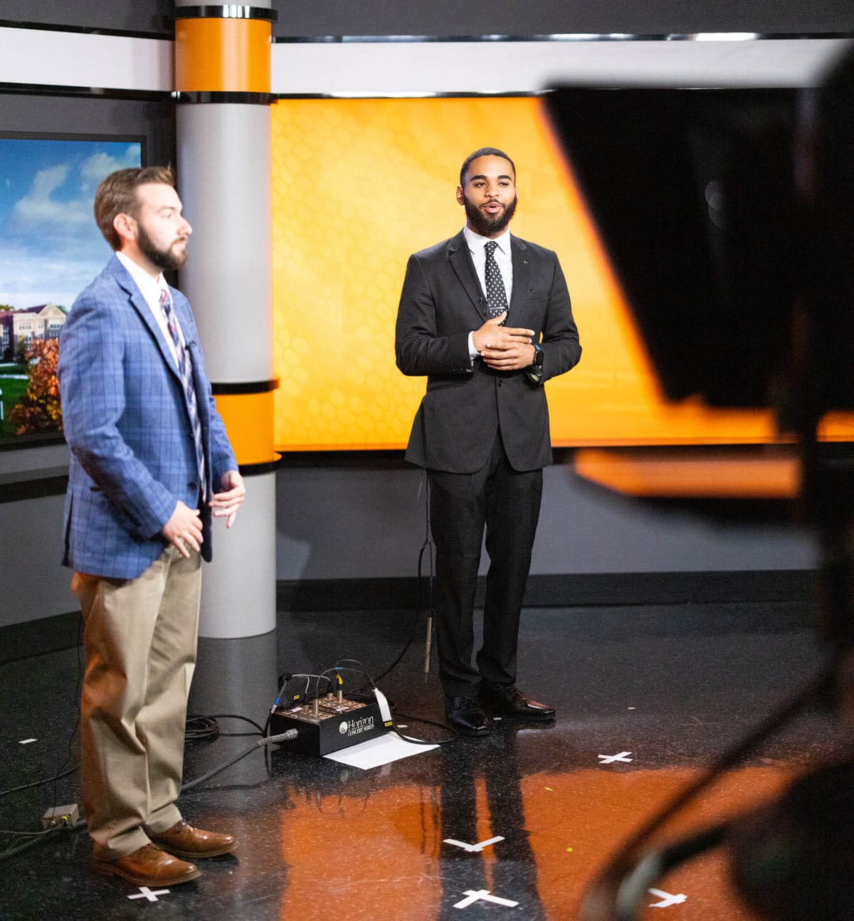 Two male students in suits get prepared to deliver a newscast at the Volunteer Channel studio.