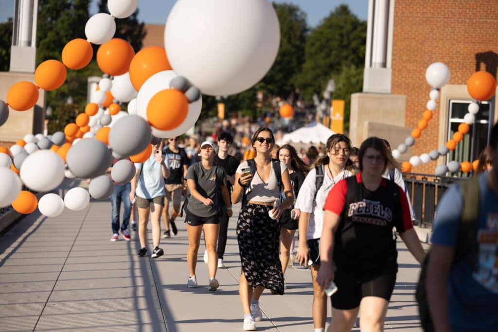 A crowd of UT students walk across the pedestrian walkway on the first day of classes, with orange, white, and grey balloon decorations floating next to them.