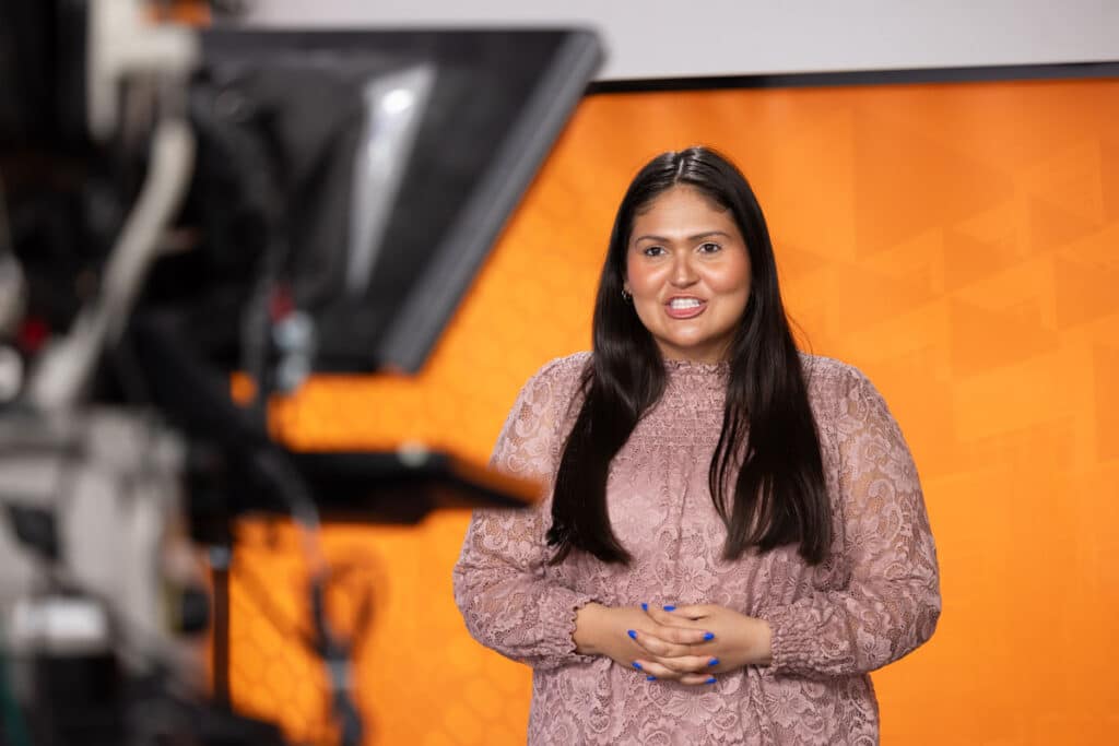 A student stands in front of a camera and with the UT orange background of the Volunteer Channel behind her as she does a broadcast.