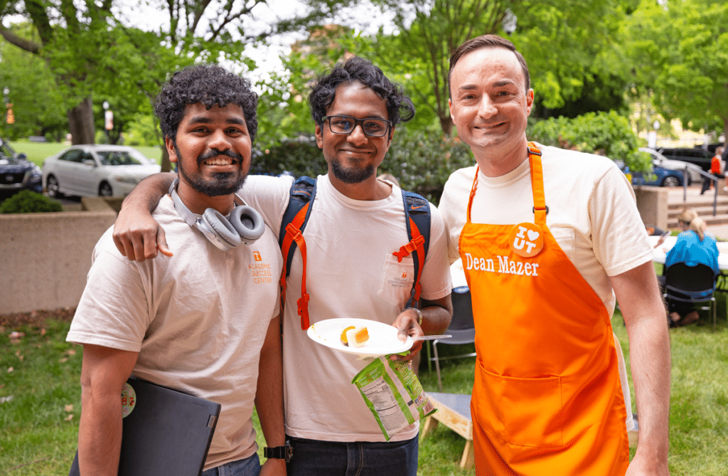 Two PhD students wearing CCI shirts pose with CCI Dean Joe Mazer, who is wearing an orange UT apron for CCI's annual Dogs with the Dean student event.