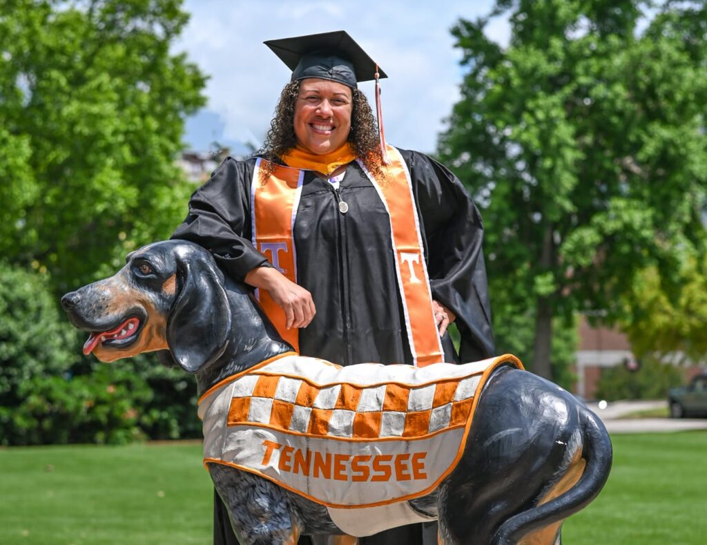 Melissa Espinales, graduate of the Master of Science in Communication and Information with a concentration in Strategic and Digital Communication program, stands in full graduation regalia behind one fo the Smokey the dog statues in circle park.