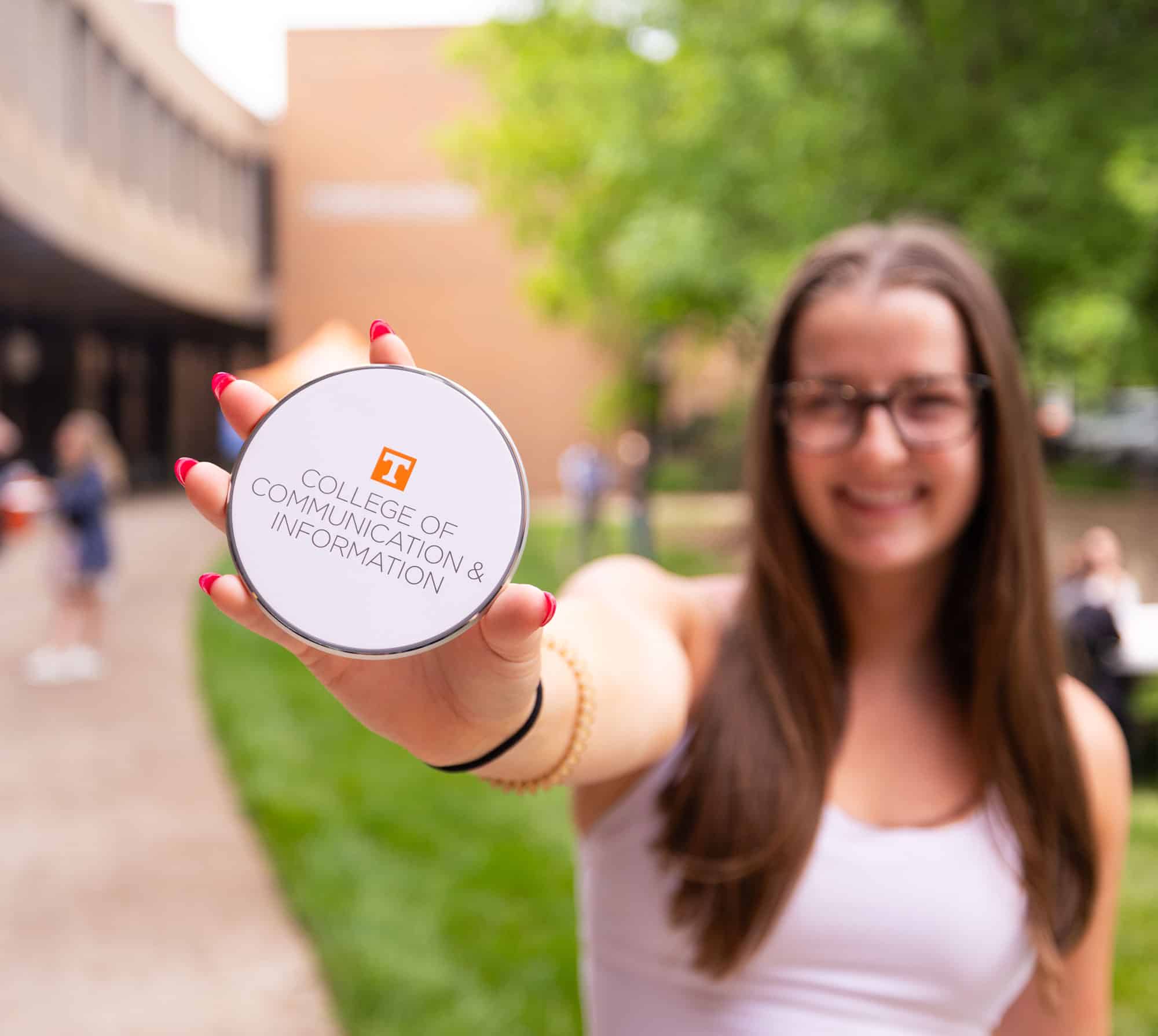 A smiling student with a hot pink manicure holds up a circular wireless charging pad with the College of Communication logo on it in the forefront. The student and background are all blurry and the charger is in focus.