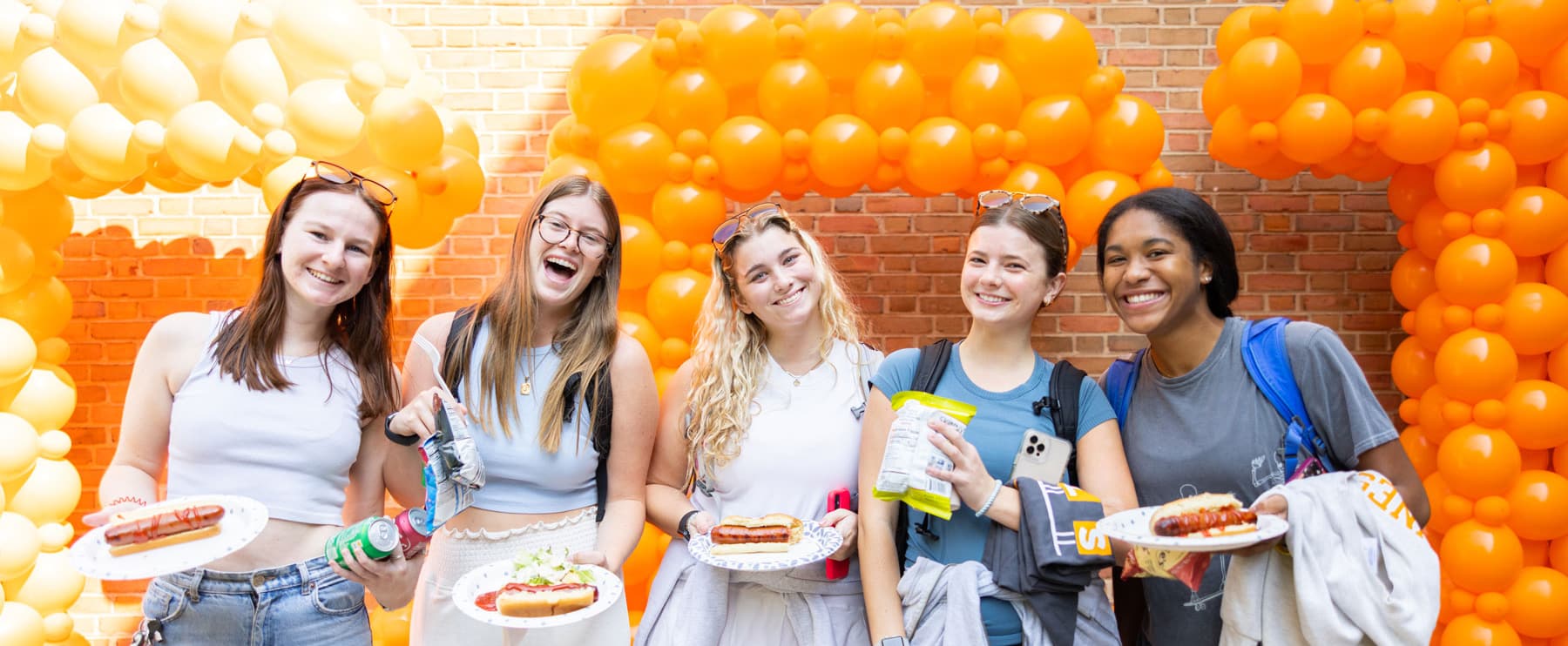 Five students stand in front of orange balloons that spell out a large CCI, all lauging and smiling and holding food from the Dogs with the Dean event.