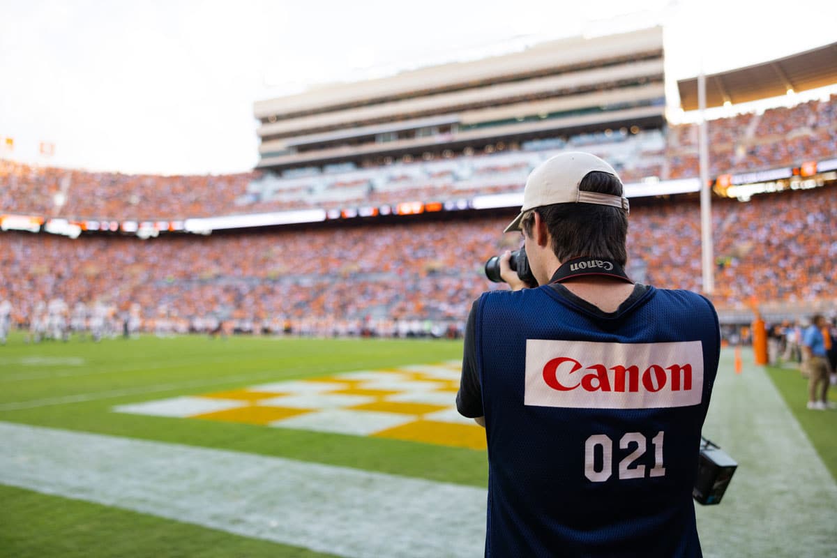 The back of a student wearing a shirt that says "Canon 021" is in the foreground while he holds a camera to his face and takes a picture of Neyland Stadium while on the field, with the field and audience-packed stadium visible.